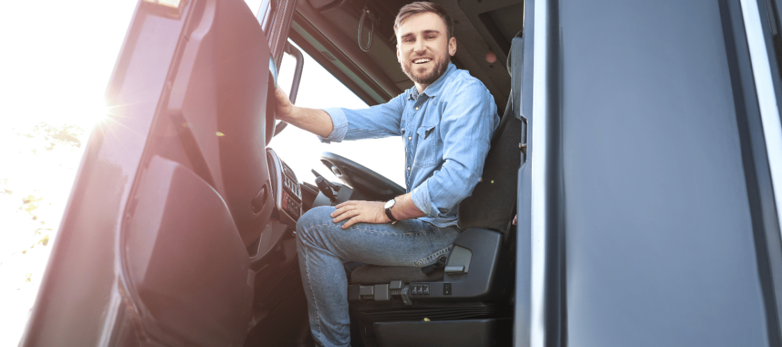 an hgv driver wearing denim sits in his cab and smiles at the camera with the vehicle door open.