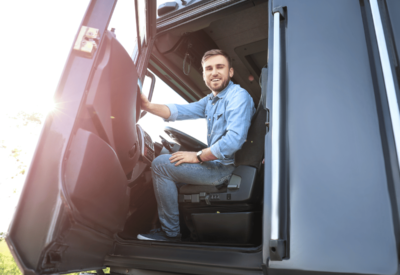 an hgv driver wearing denim sits in his cab and smiles at the camera with the vehicle door open.