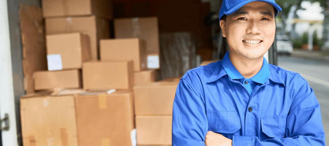 a delivery driver wearing a blue shirt and blue cap stands in front of his hgv vehicle. the back of the lorry is open and is loaded with boxes.