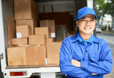 a delivery driver wearing a blue shirt and blue cap stands in front of his hgv vehicle. the back of the lorry is open and is loaded with boxes.