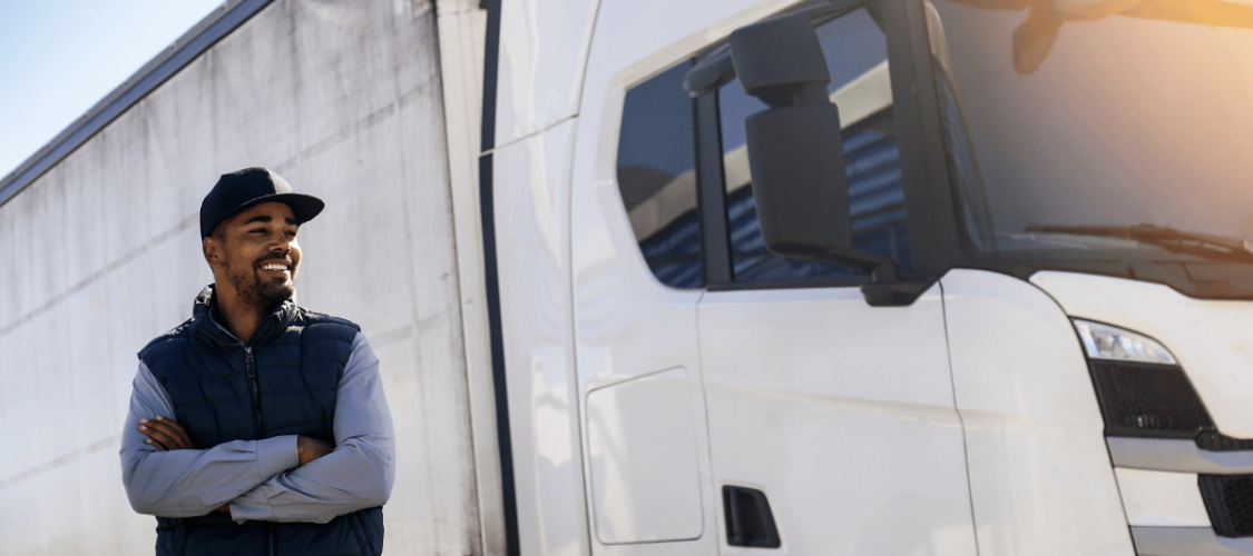 a man wearing a gillet and baseball cap stands next to a white hgv. he is smiling after completing hgv driver training.