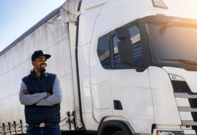 a man wearing a gillet and baseball cap stands next to a white hgv. he is smiling after completing hgv driver training.